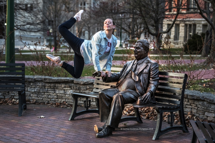 Person leaps in the air leaning on the arm of the statue of Herman B Wells. Their legs are bent at the knees midair to their side.