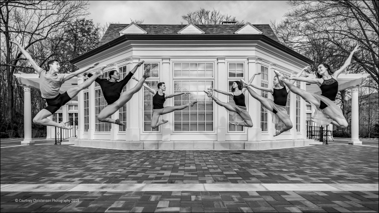 Six dancers jump in the air with one leg extended and toes pointed with their other leg bent underneath them. They jump in front of a building with floor to ceiling windows.