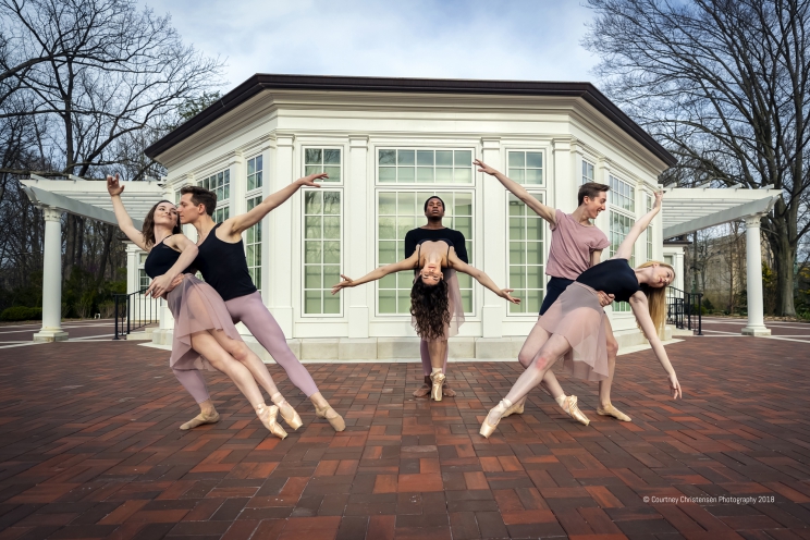 Six dancers coupled together pose with their arms extended in front of a white building.