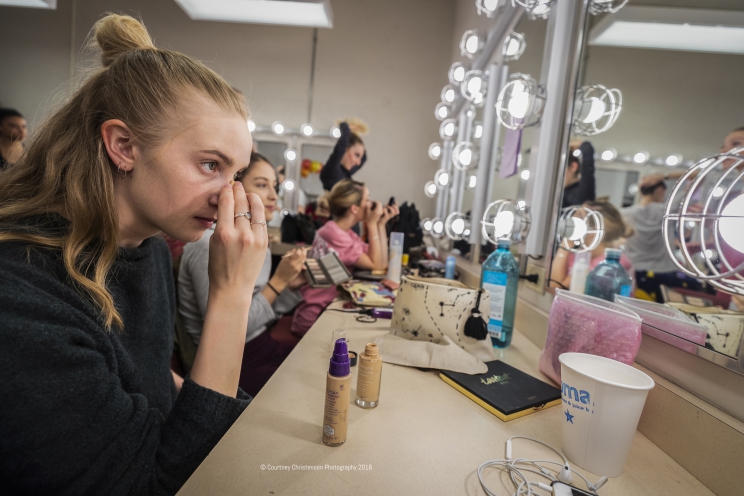Several people sit and apply makeup in front of mirrors with lights surrounding them.
