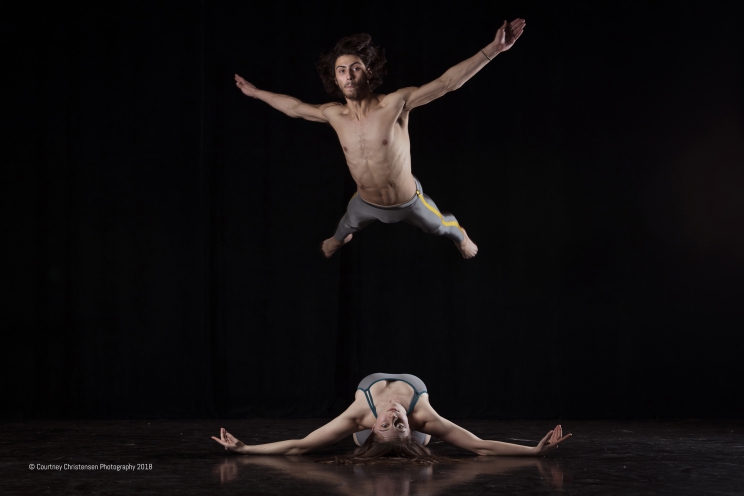 Person lays on their back with their arms extended and head looking back at the camera as another person jumps above them with their arms and legs extended.