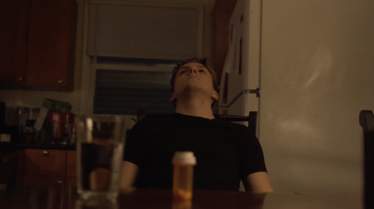 A man sits at a table and looks at the ceiling. A glass of water and pill bottle sit in front of him.
