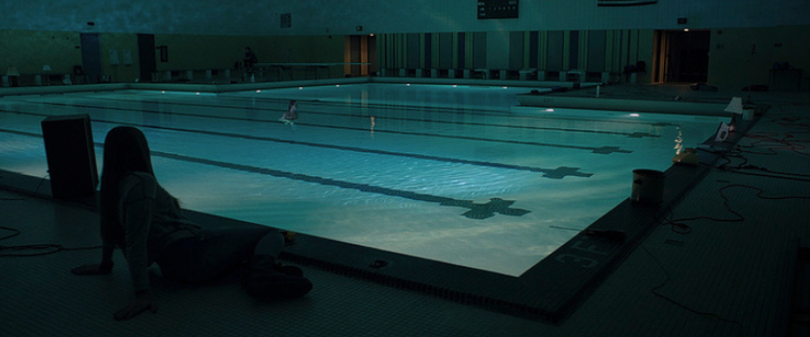 A woman swims alone in an indoor pool. The lights are off. Another woman sits on the side of the pool.