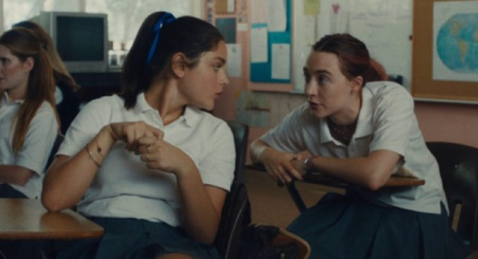 Two girls in uniform talk over school desks.